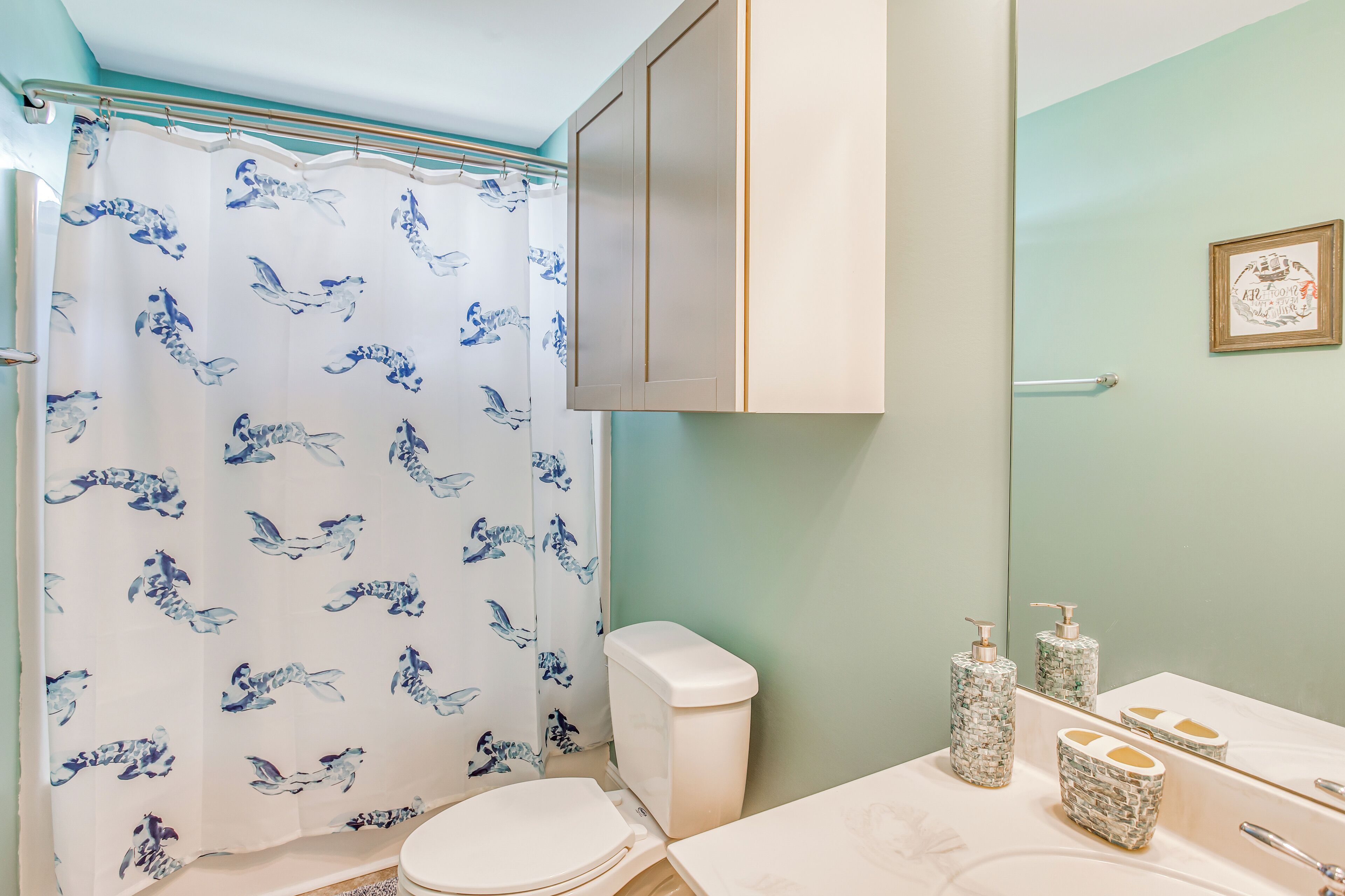 A bathroom with a toilet, a shower curtain with a blue pattern, a sink with a mirror, and a cabinet above the sink.