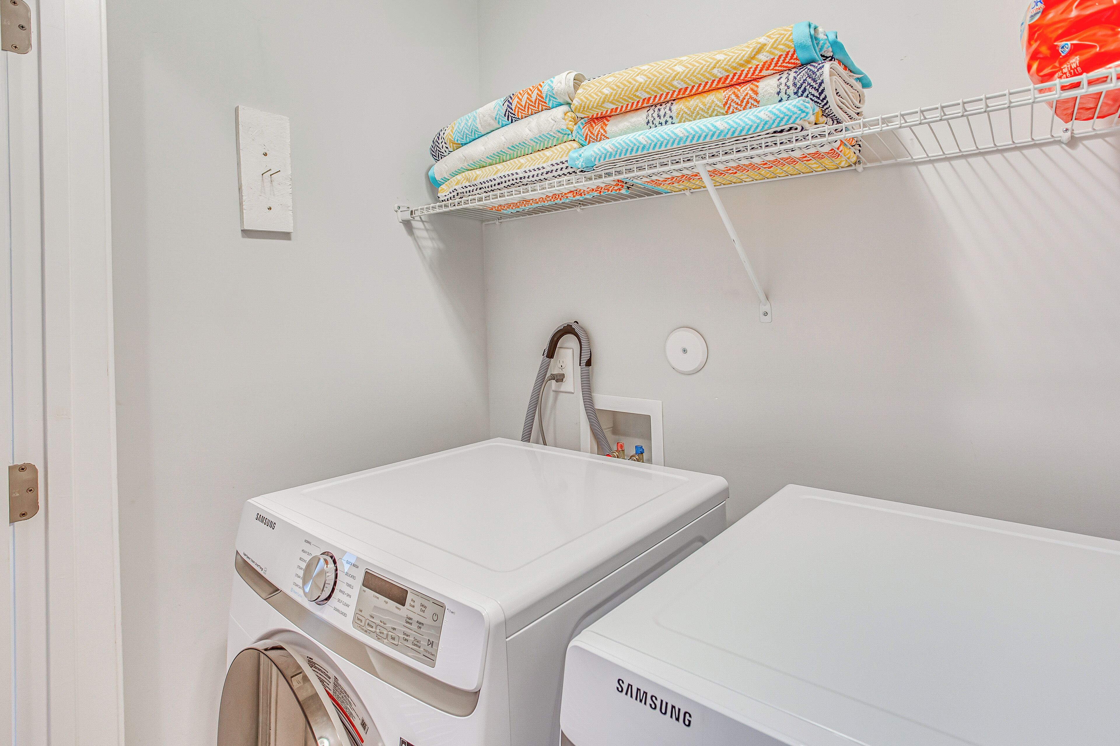 A laundry room with two white Samsung washing machines, a metal drying rack with colorful towels, and a white door.