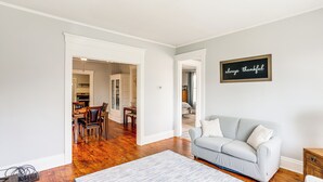 Ein Wohnzimmer mit grauem Sofa, einem Couchtisch und einem Teppich. Es gibt einen Essbereich mit Tisch und Stühlen. Ein Schild an der Wand zeigt den Schriftzug 'always thankful'.