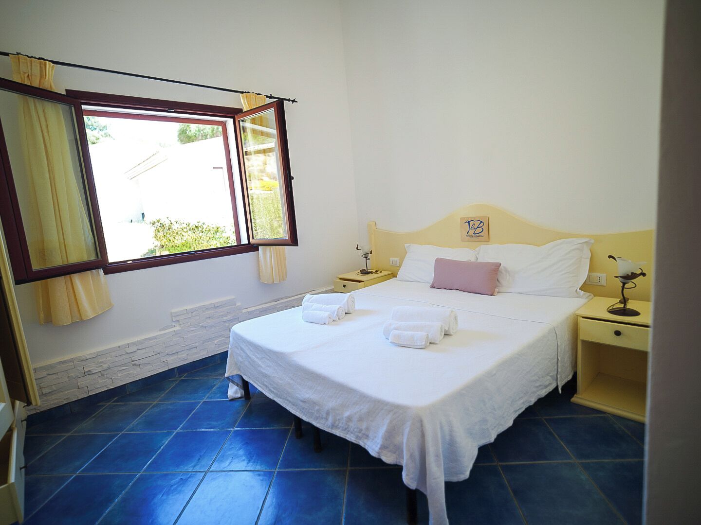 A bedroom with a bed, two bedside tables, a window with curtains, and a stone wall.