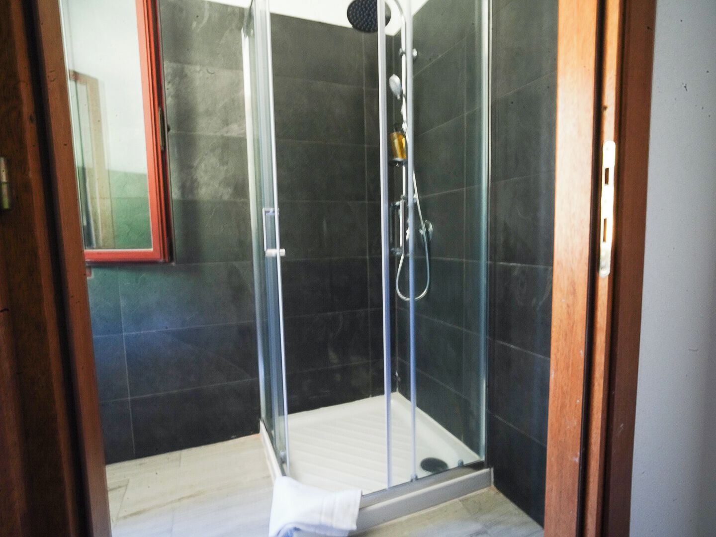 Cabina de ducha con paredes de vidrio, piso blanco, cabezal de ducha y un rociador de mano.
