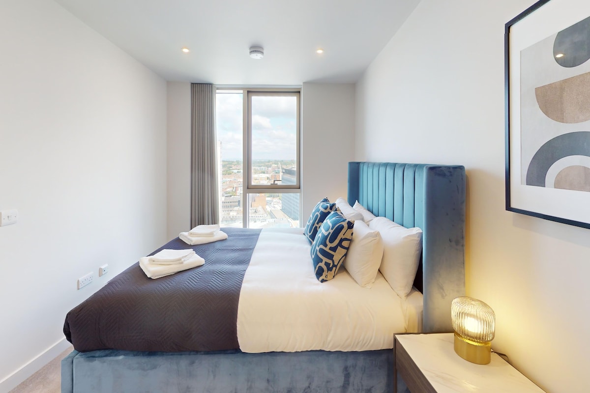 A modern hotel room with a large bed, a view of the city, and a framed abstract artwork on the wall.