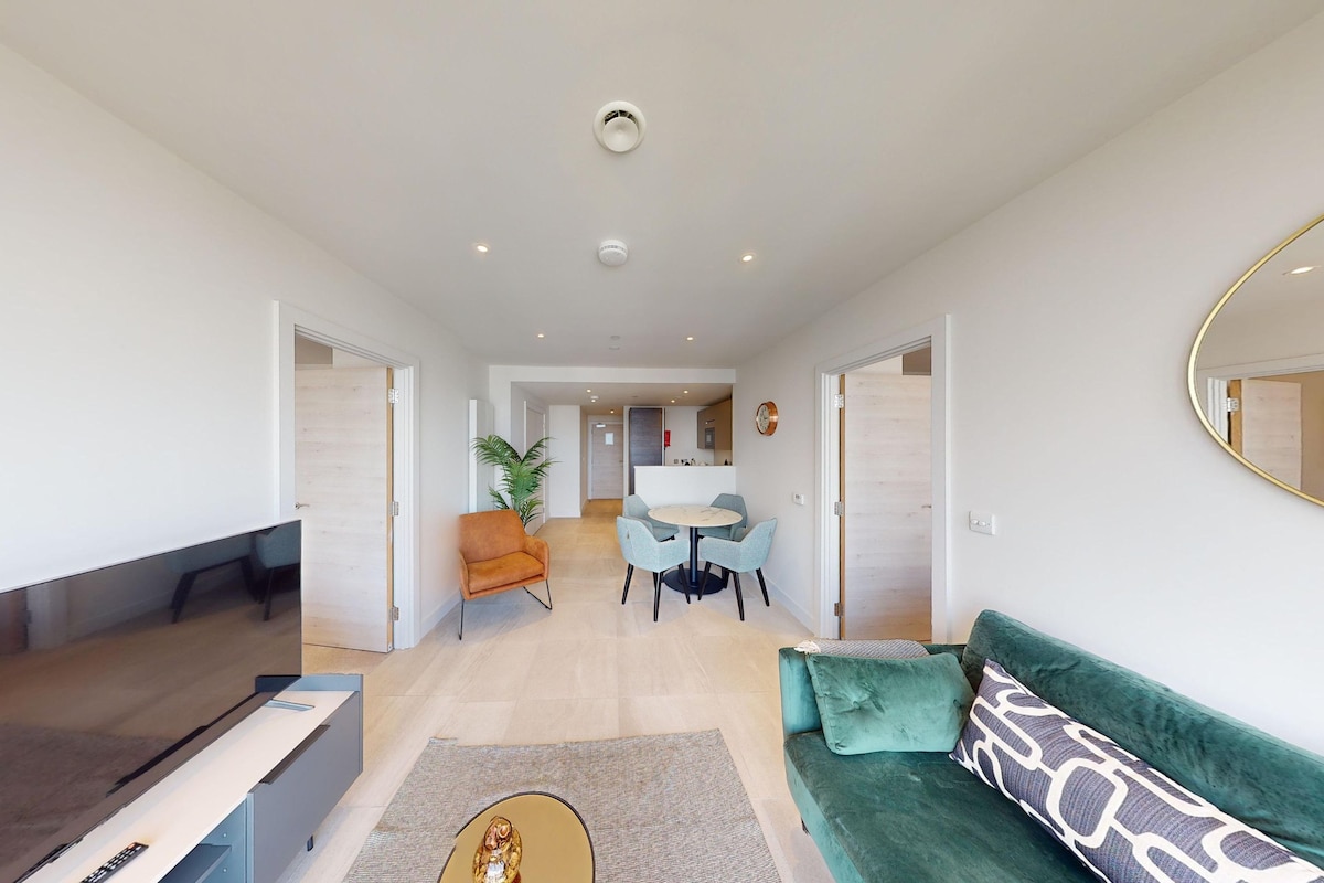 A modern living room with a sofa, a coffee table, and a TV. There is a dining area with a table and chairs. The room has a ceiling light and a door leading to another room.