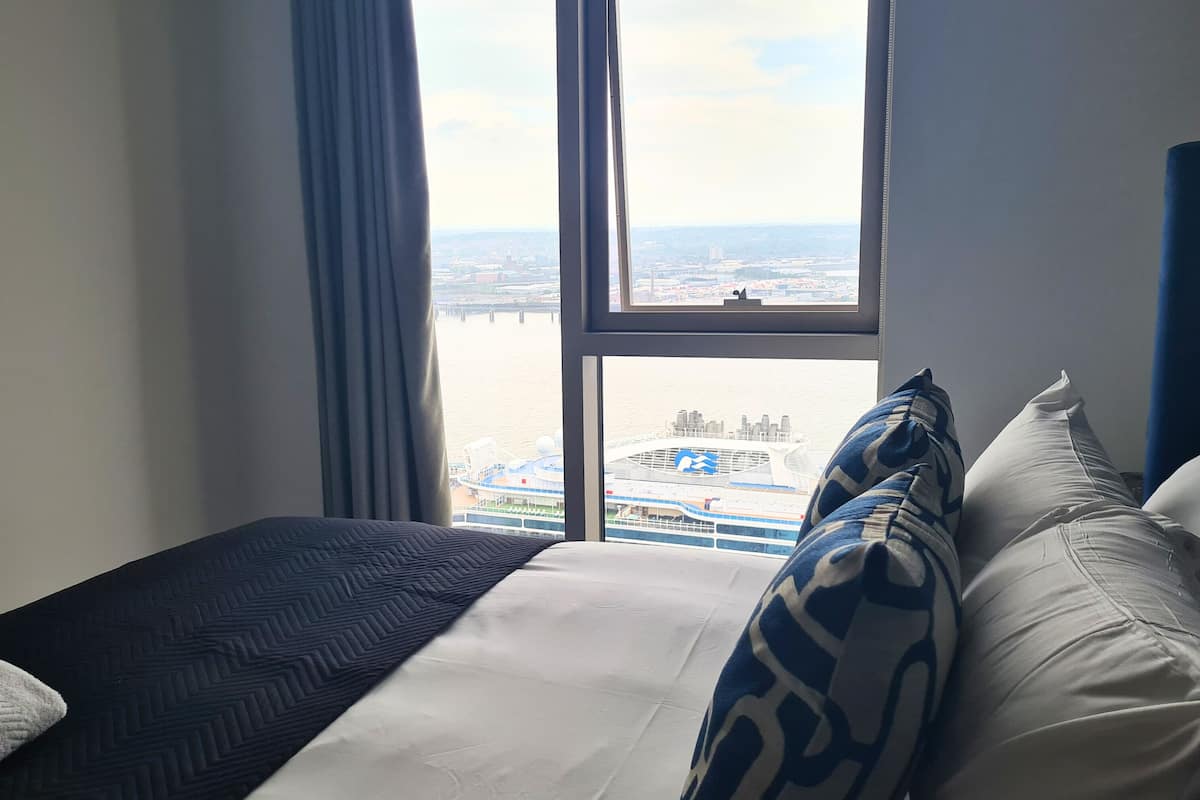 A hotel room with a large window, a bed with white and patterned pillows, and a view of a cruise ship docked at a port.