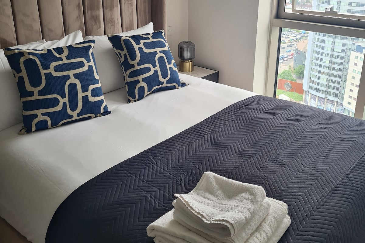 A neatly made bed with a dark quilt and two patterned pillows, a bedside lamp, and a view of a cityscape through a large window.