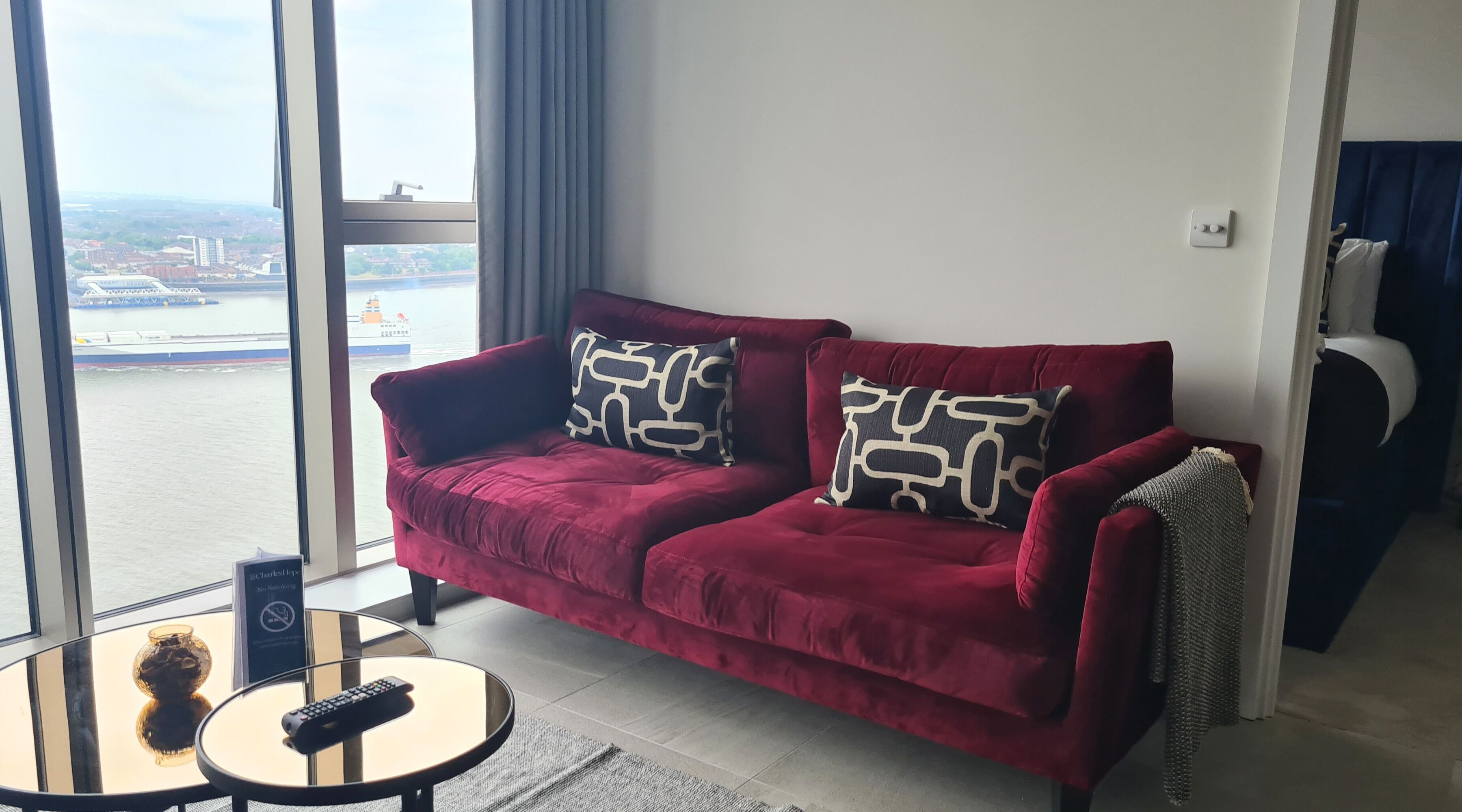 A red velvet sofa with patterned cushions, a glass coffee table with a remote control, and a view of a body of water with boats.