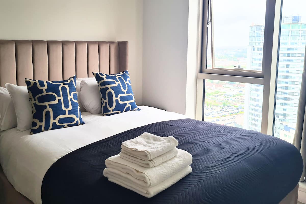 A hotel room with a bed, two pillows, a bedside table, and a window with a city view.