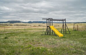 Ein Spielplatz mit Rutsche und Schaukeln in einer grünen Wiese, im Hintergrund Berge.