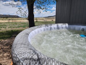 Un bain à remous avec une structure extérieure en bois, entouré d’un champ verdoyant et d’un arbre.