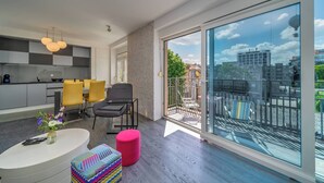 A modern living room with a large sliding glass door leading to a balcony, a kitchen area with grey cabinets, and a dining set. - Silver Apartment With Hot Tub And Balcony (Zagreb)