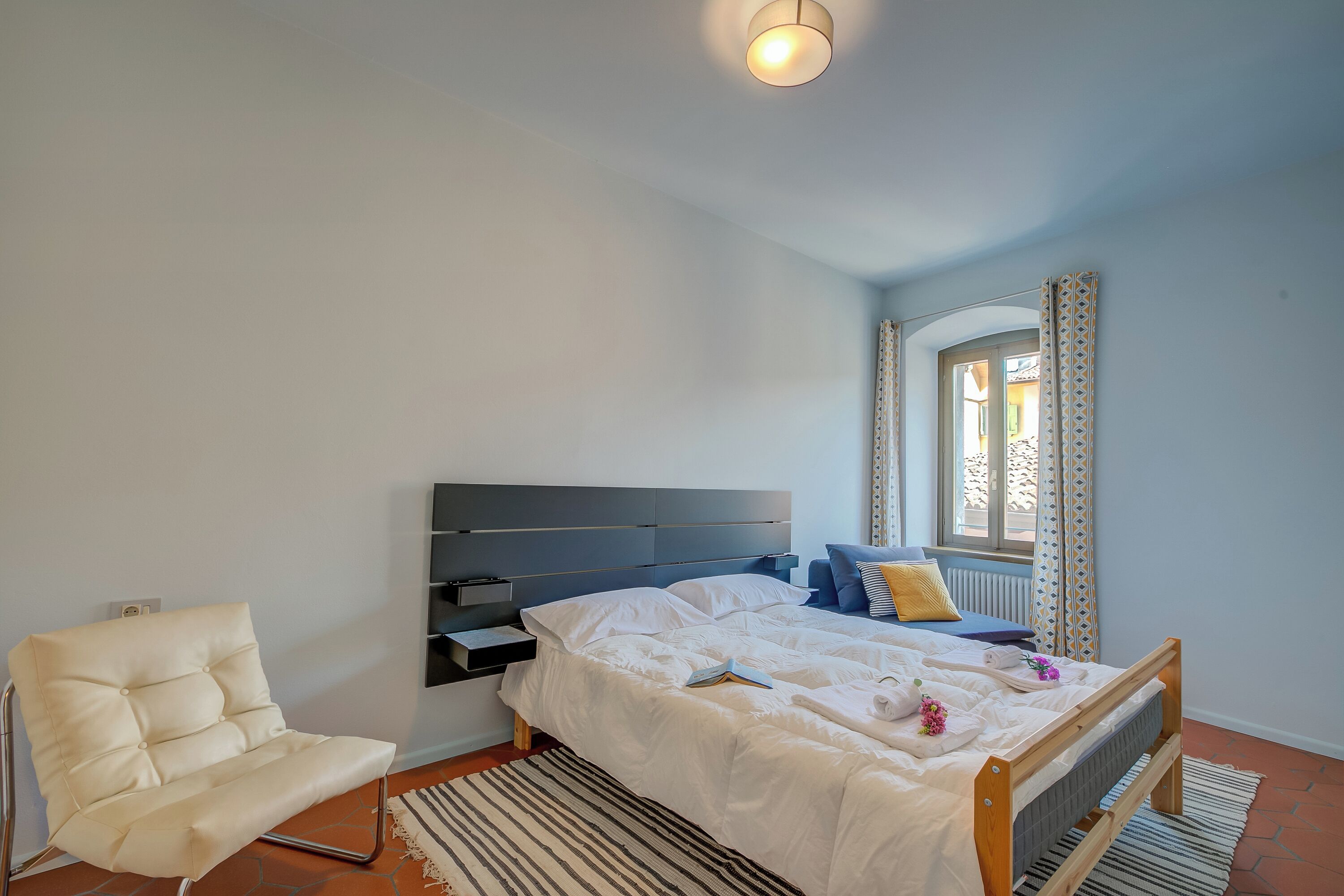 Une chambre à coucher comprenant un lit, une chaise, une table de chevet, une fenêtre avec des rideaux et un radiateur.