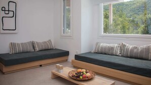 Una habitación moderna con dos sofás verdes, una mesa de centro de madera con una cesta de frutas y un gran ventanal con vistas a un bosque.