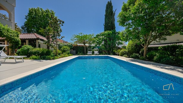 Une piscine d’un bleu éclatant, entourée d’une végétation luxuriante, et un bâtiment surmonté d’un toit en tuiles.