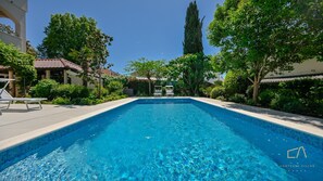 Une piscine d’un bleu éclatant, entourée d’une végétation luxuriante, et un bâtiment surmonté d’un toit en tuiles.