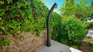 Une douchette d’extérieur noire, fixée à un mur de pierre, est entourée d’un feuillage verdoyant et luxuriant.