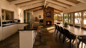 A modern kitchen with a central island, a stone fireplace, and a dining area with chairs. - Glasshouse Lake Eildon Architectural Retreat (Taylor Bay)