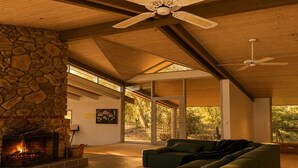A modern living room with a stone fireplace, green sofas, a wooden coffee table, and a ceiling fan. - Glasshouse Lake Eildon Architectural Retreat (Taylor Bay)