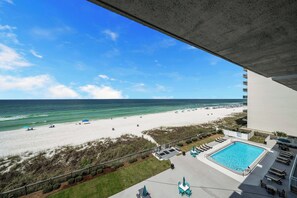 A beach view from a balcony overlooking a swimming pool with lounge chairs and umbrellas.