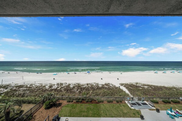 A beach view from a balcony with a clear sky, a sandy beach, and a fenced area with grass and shrubs.