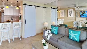 Una sala moderna con un sofá gris, almohadas turquesas y una mesa de centro. Hay dos taburetes blancos y una cocina con armarios blancos y lámparas colgantes amarillas.