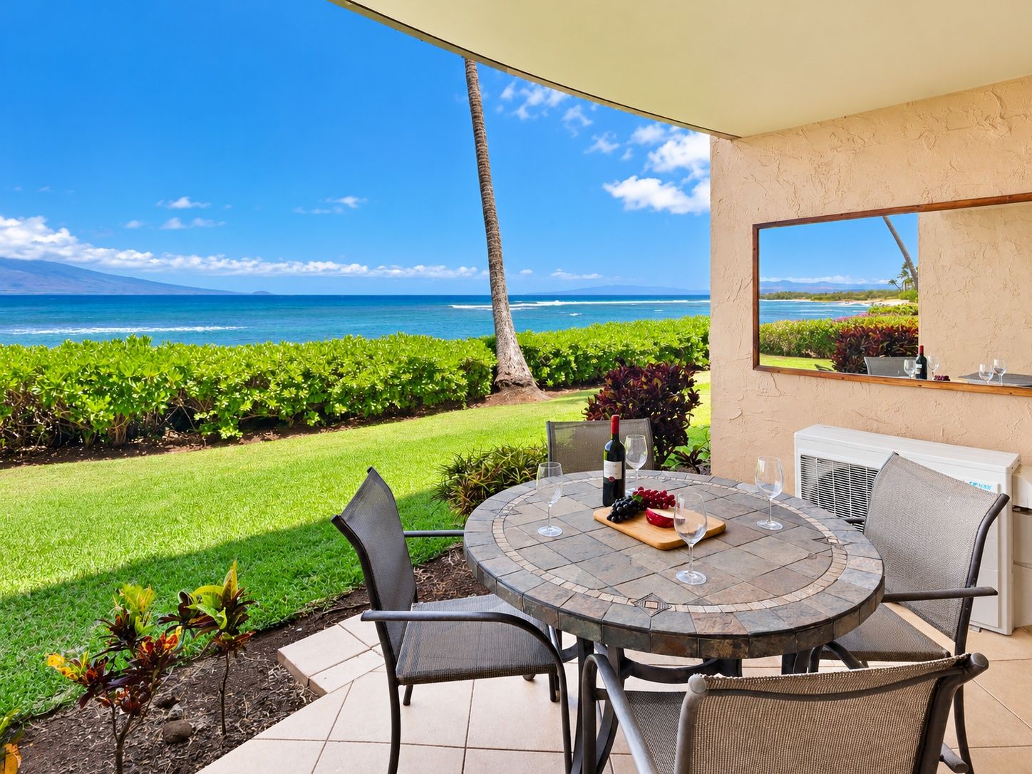 A round table with chairs, a bottle of wine, and a bowl of fruit on a patio overlooking a lush garden and the ocean.