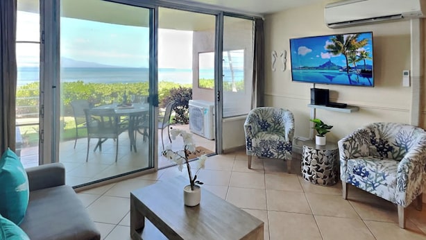 Una sala de estar moderna con un gran ventanal corredero que da a un balcón con vistas al océano. Hay un televisor de pantalla plana montado en la pared, una mesa de centro y dos sillones con estampado.