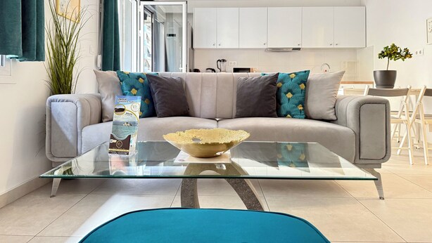 Un salon moderne avec une table basse en verre, un canapé agrémenté de coussins à motifs et, au fond, une vue sur la cuisine.