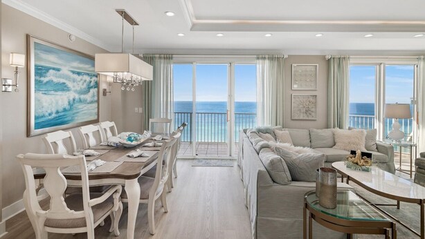 A spacious living room with a large window overlooking the ocean, a dining area with a white table and chairs, and a cozy seating area with a sofa and armchairs.