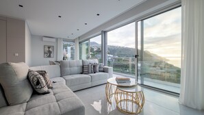 A modern living room with a large sectional sofa, a round coffee table, and a view of a coastal landscape through floor-to-ceiling windows. - Luxury Villa Pérola (Calheta)