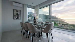 A modern dining area with a glass table and chairs, a painting on the wall, and a view of a coastal landscape through large windows. - Luxury Villa Pérola (Calheta)