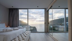 A modern bedroom with a large bed, a bench, and a view of the sea and hills. - Luxury Villa Pérola (Calheta)