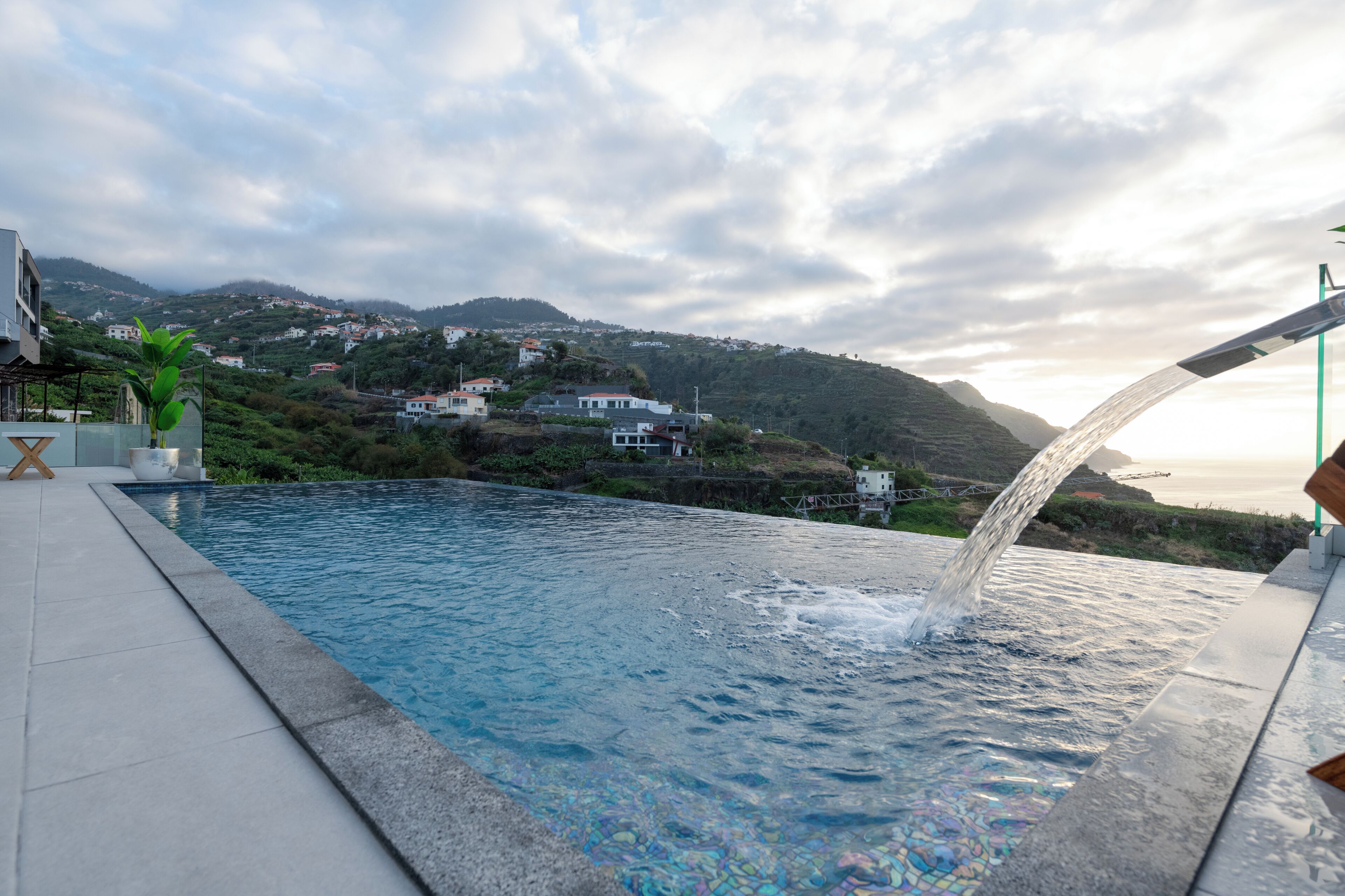Une piscine dans laquelle l’eau s’écoule, entourée d’une terrasse carrelée et offrant une vue sur une colline parsemée de maisons.