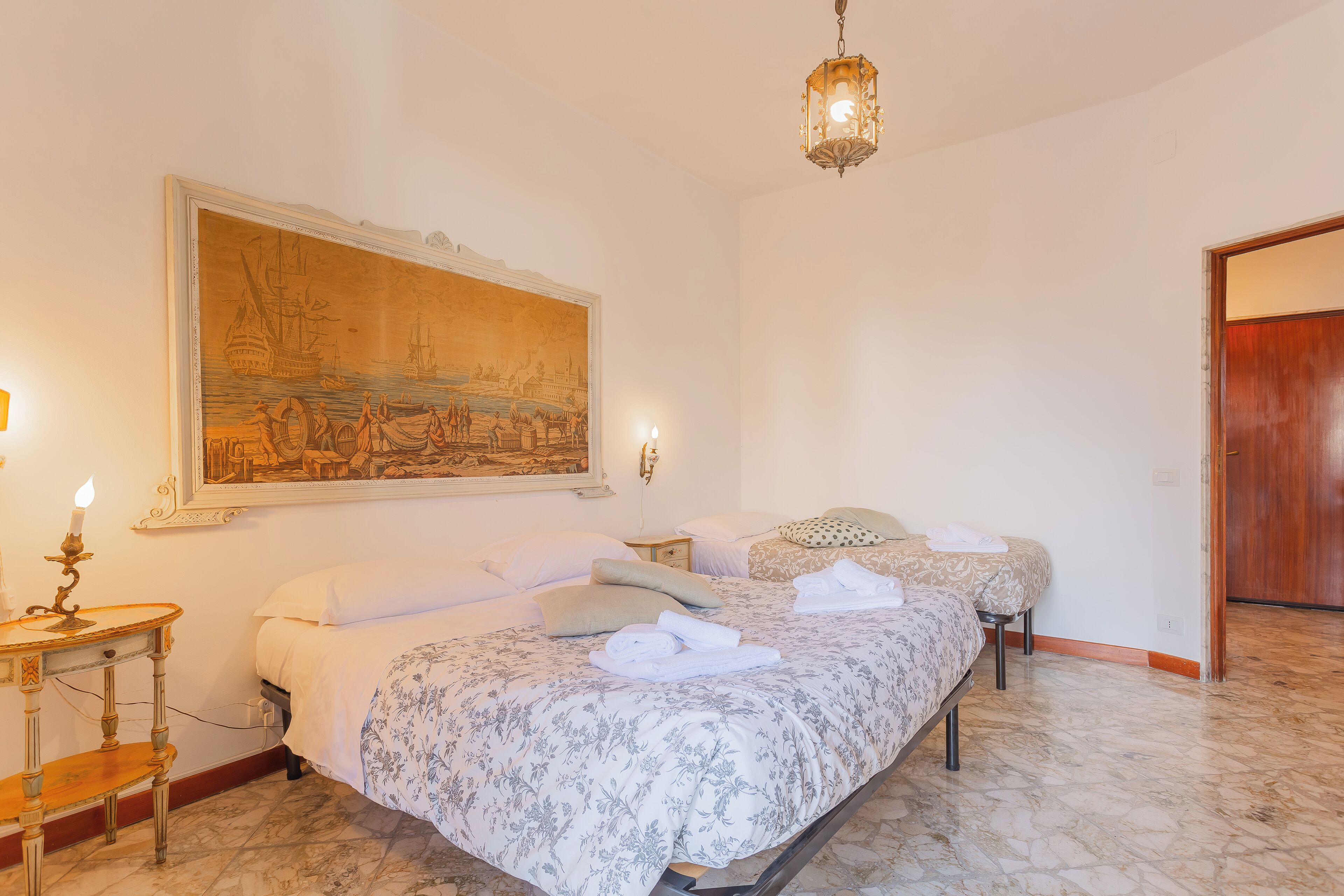 A bedroom with two beds, a large painting on the wall, a wooden side table with a candle, and a hanging light fixture.