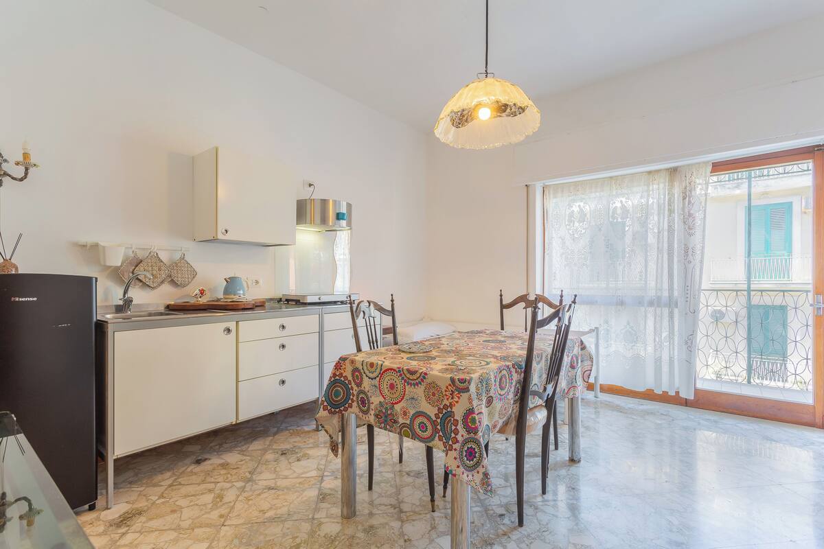 Un coin repas avec une table recouverte d’une nappe à motifs, un luminaire suspendu et une vue sur un balcon doté d’une balustrade.