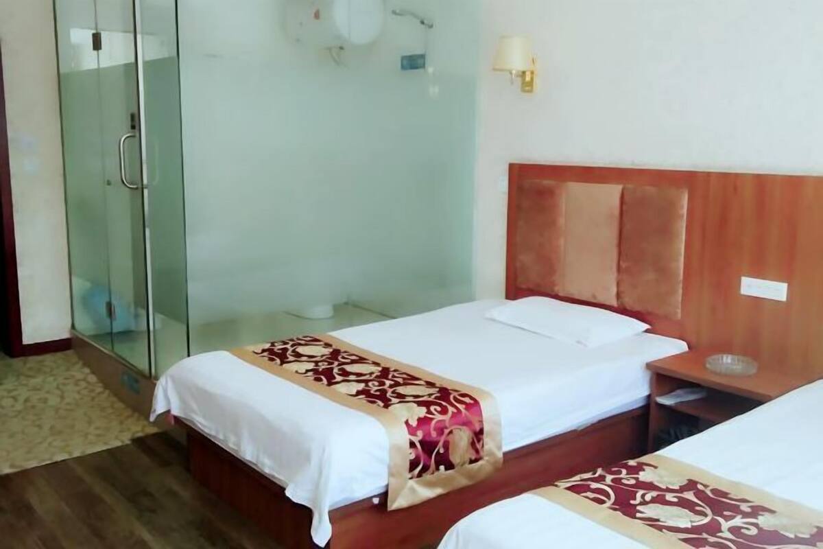 A hotel room with two beds, a wooden headboard, a glass shower enclosure, and a wall-mounted lamp.
