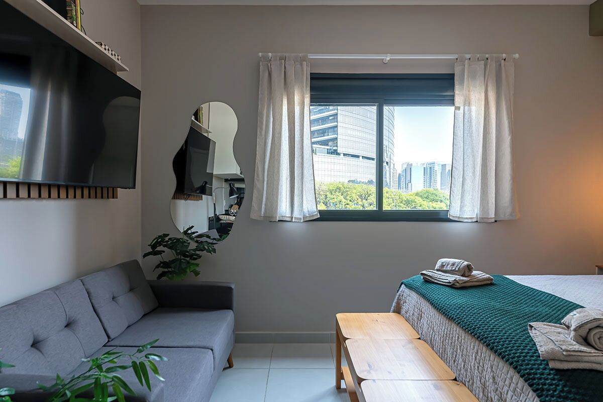 Una habitación moderna con sofá, cama, banca de madera, espejo y ventana con cortinas.