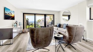 A modern living room with a sofa, two armchairs, a glass coffee table, and a dining table. There is a large window with a view of the outdoors.