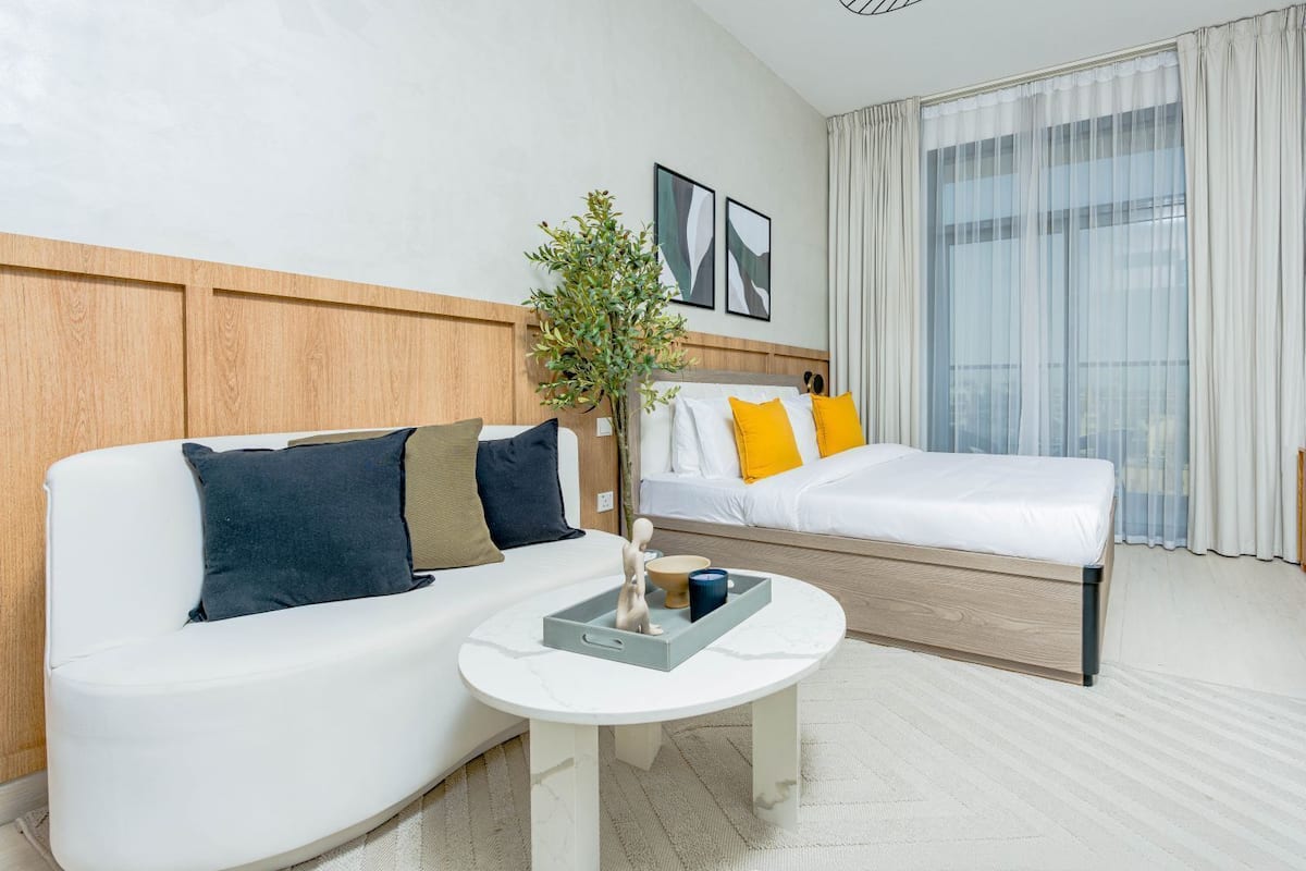 A hotel room with a white sofa, a bed with yellow pillows, a small table with a tray, and a large window with sheer curtains.