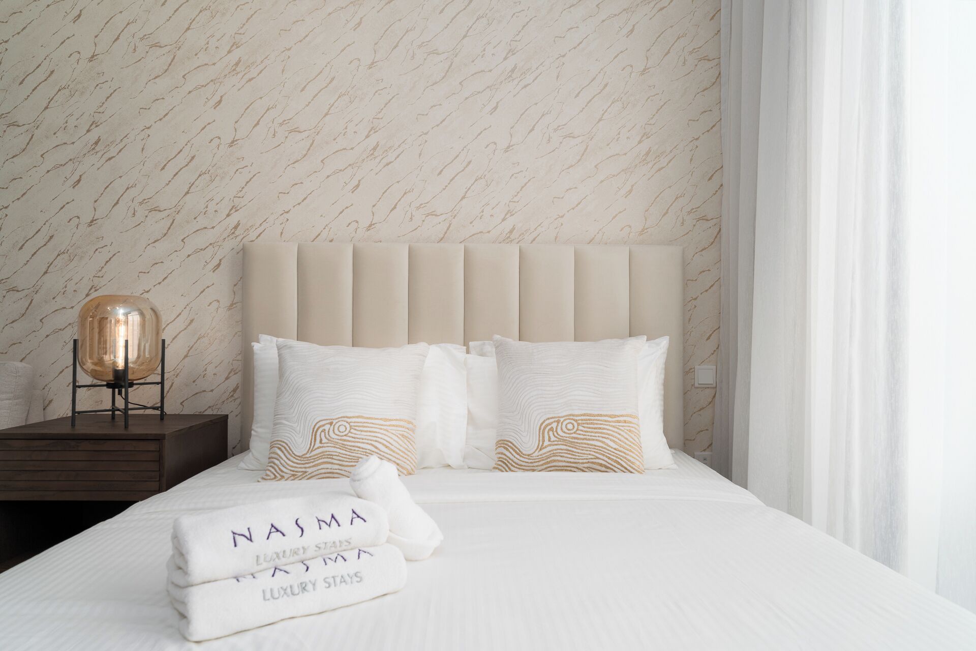 A neatly made bed with white linens and two decorative pillows, a bedside table with a lamp, and a marble-patterned wall.