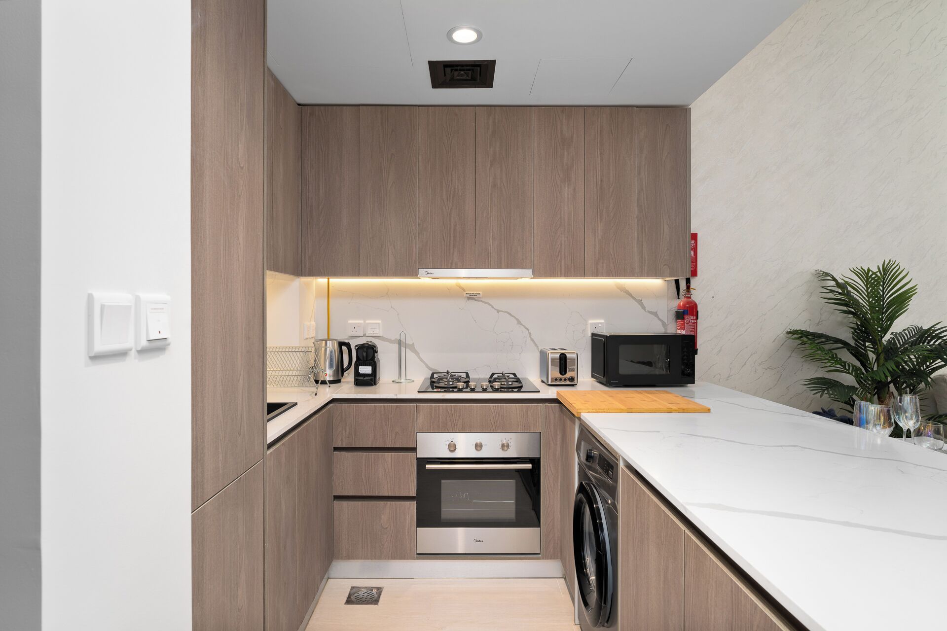 Cocina compacta con horno empotrado, microondas y lavavajillas, armarios de madera y encimera blanca.