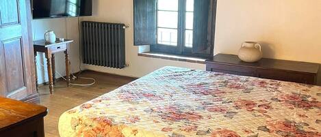 A bedroom with a wooden ceiling, a bed with a floral quilt, a radiator, a window with shutters, and a television on the wall.