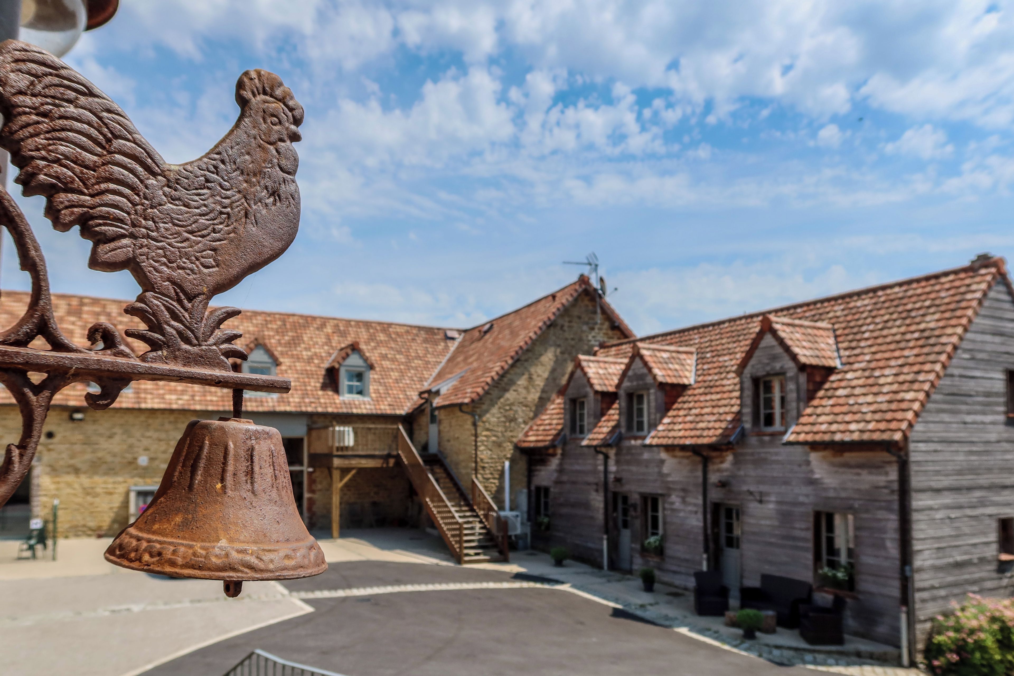 Une cour bordée de bâtiments traditionnels, ornée d’une sculpture de coq et d’une cloche.