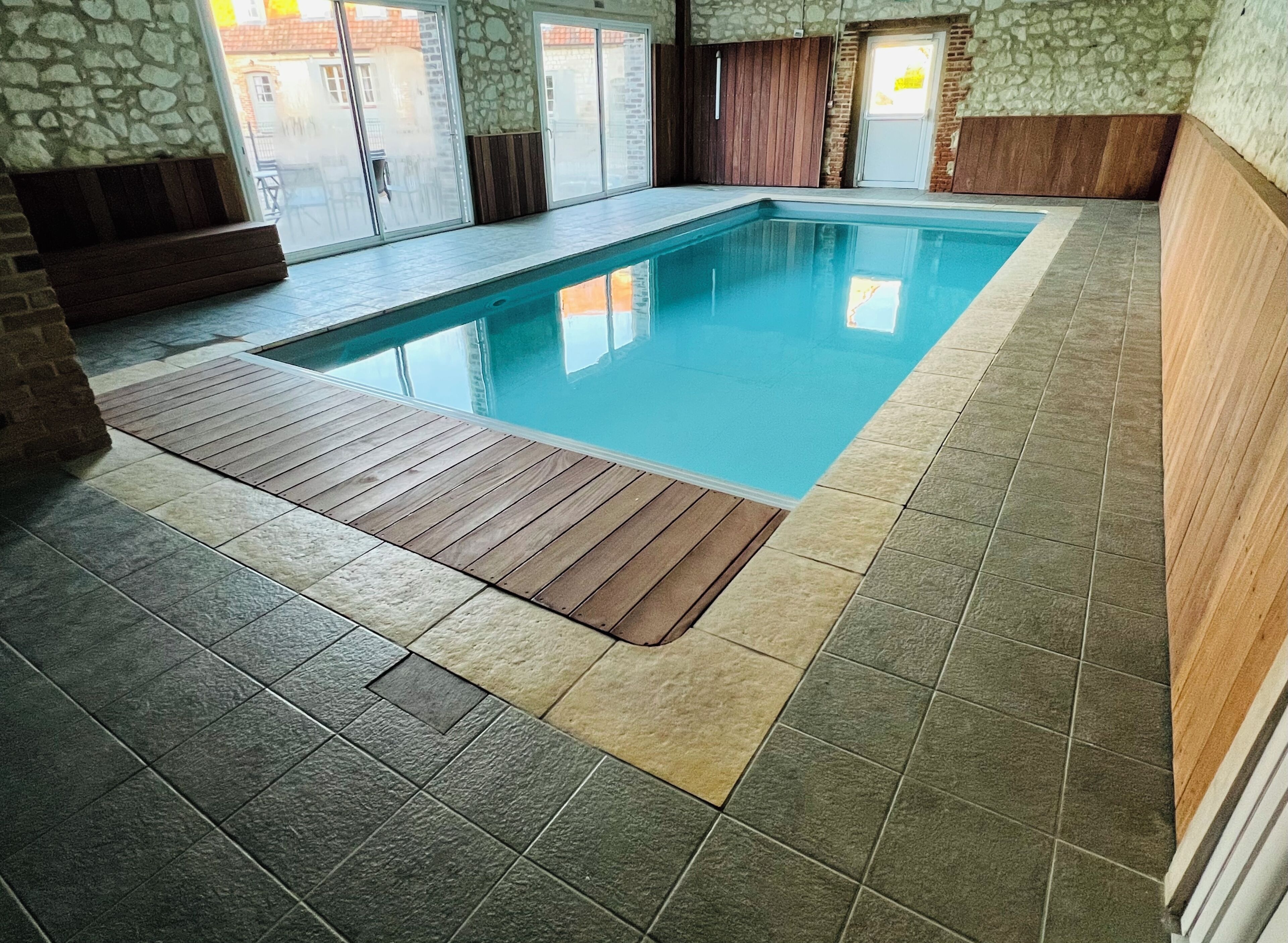 An indoor swimming pool with a wooden deck, stone walls, and a tiled floor.