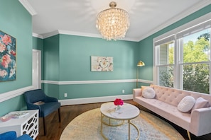 Et værelse med teal vægge, hvidt loft, en lysekrone, en rosa sofa, en blå stol, et rundt bord med en vase og et stort vindue.