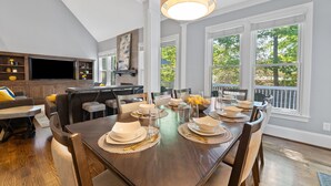 A dining area with a wooden table set for six, surrounded by chairs, with a kitchen and living area in the background. - Spacious 5 Bedroom, Hot Tub, Arcade Room, EV Charger, Handicap & Quiet Culdesac! (Raleigh)