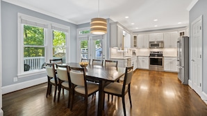 A modern kitchen with white cabinetry, a dining area with a wooden table and chairs, and a large window with a view of greenery. - Spacious 5 Bedroom, Hot Tub, Arcade Room, EV Charger, Handicap & Quiet Culdesac! (Raleigh)
