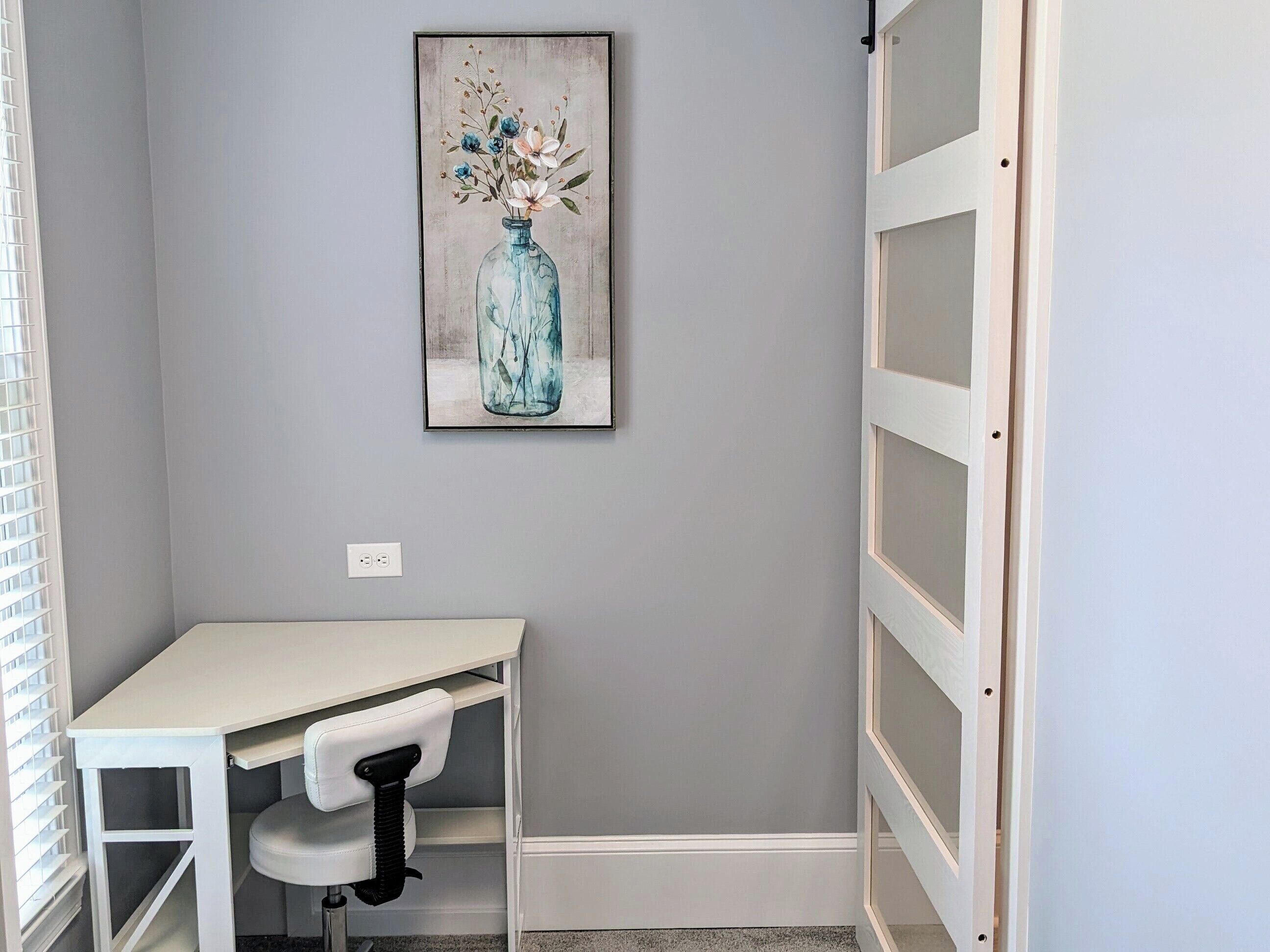 Una habitación pequeña con un escritorio blanco, una silla y un cuadro de un jarrón azul con flores en la pared.