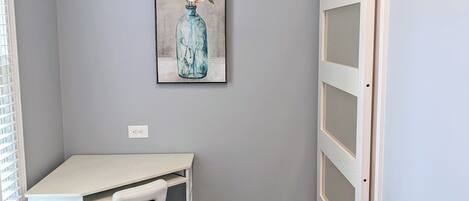 Una habitación pequeña con un escritorio blanco, una silla y un cuadro de un jarrón azul con flores en la pared.