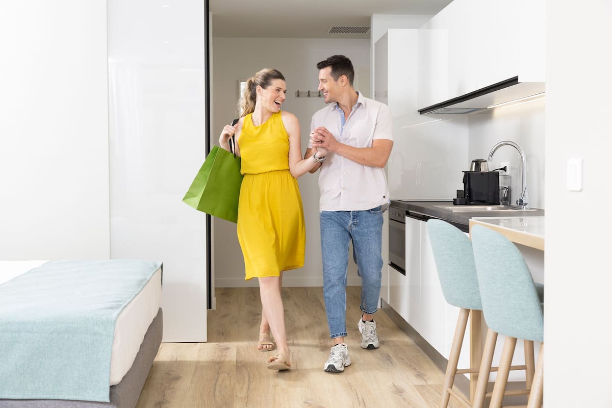 A modern kitchen with white cabinets, a black countertop, and a stainless steel sink. There are two bar stools with light blue upholstery.