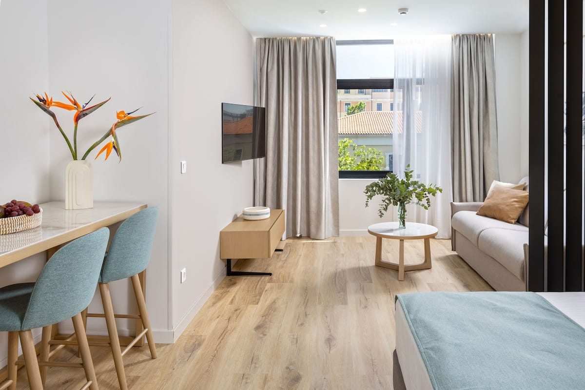 A modern hotel room with a wooden floor, a desk with chairs, a sofa, a bed, a small table with a plant, and a TV on the wall.